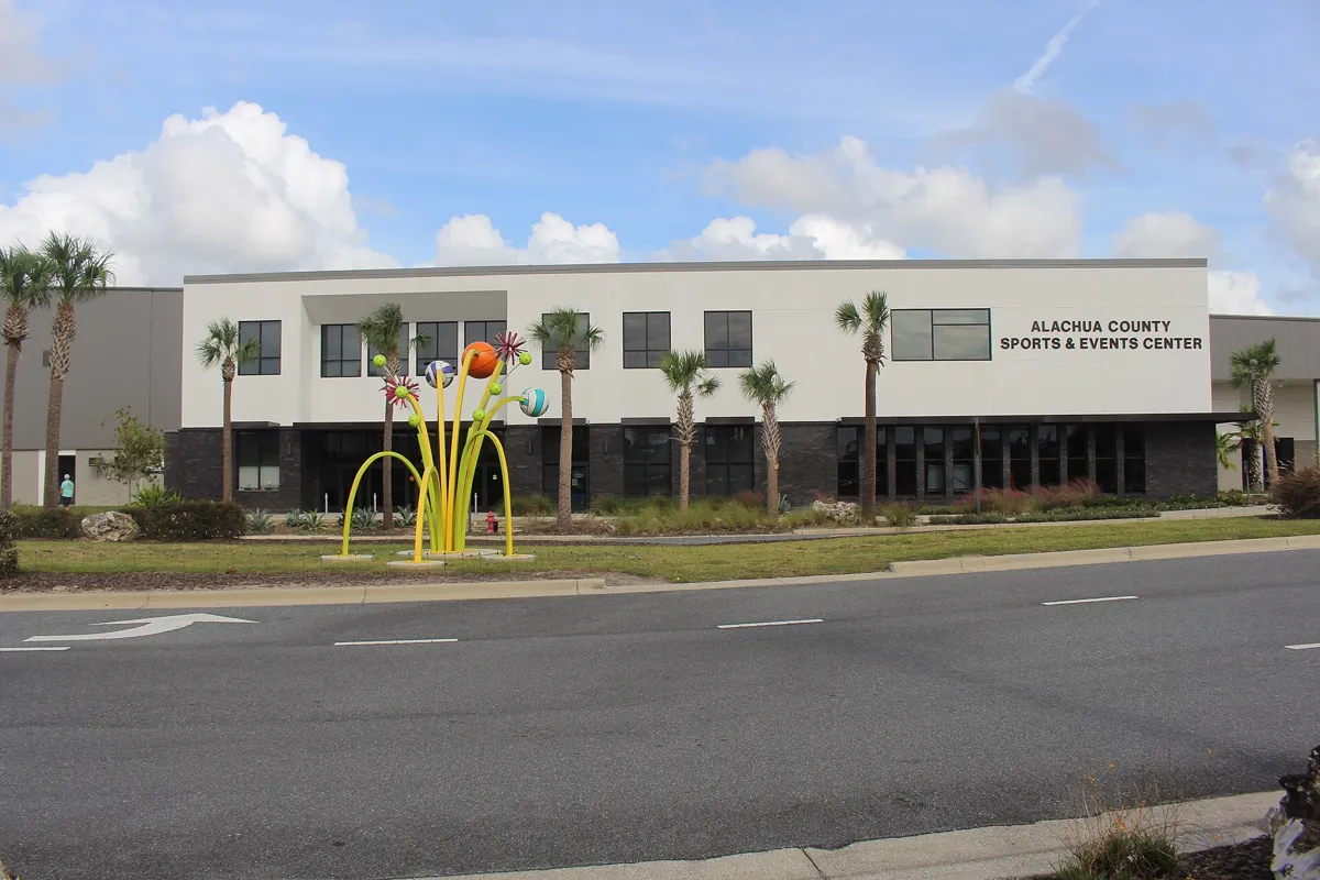 The front of the Alachua County Sports and Events Center, a white two-story building with palm trees and a yellow sculpture of sports balls outside.